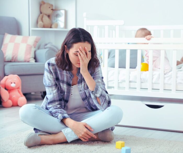 A young mother sitting on the floor in front of her baby's crib and feeling sad and overwhelmed