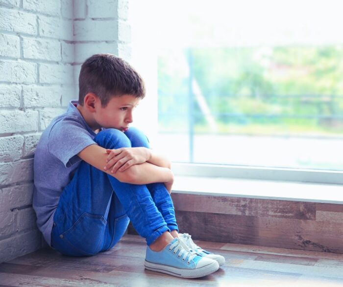 A little boy sitting on the floor in front of a window looks very sad