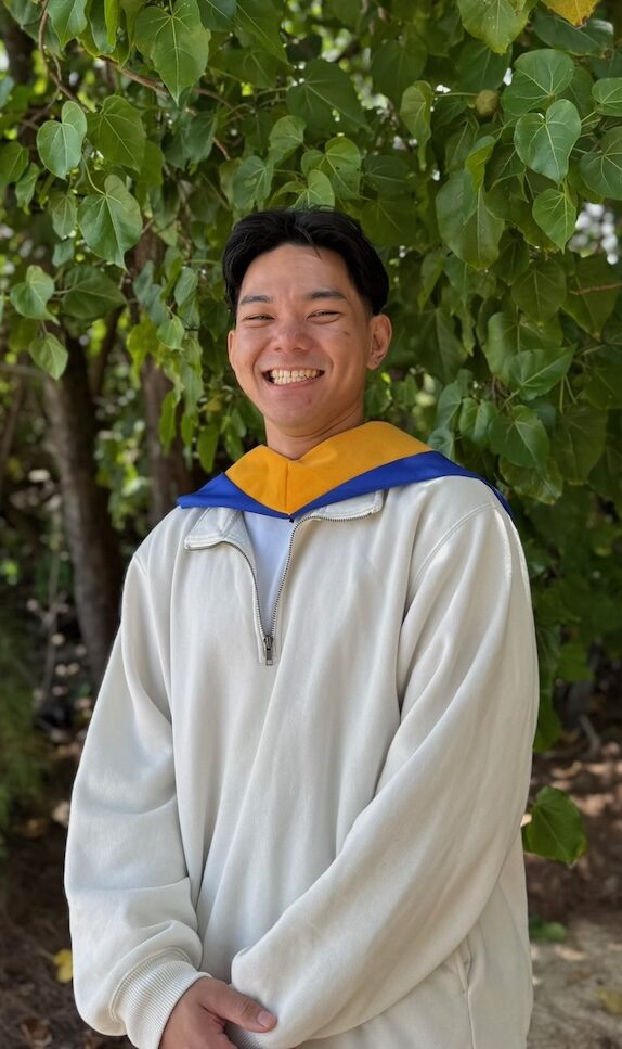 Smiling young man wearing a light-colored hoodie with a blue and orange hood standing outdoors among green leaves, looking at the camera.