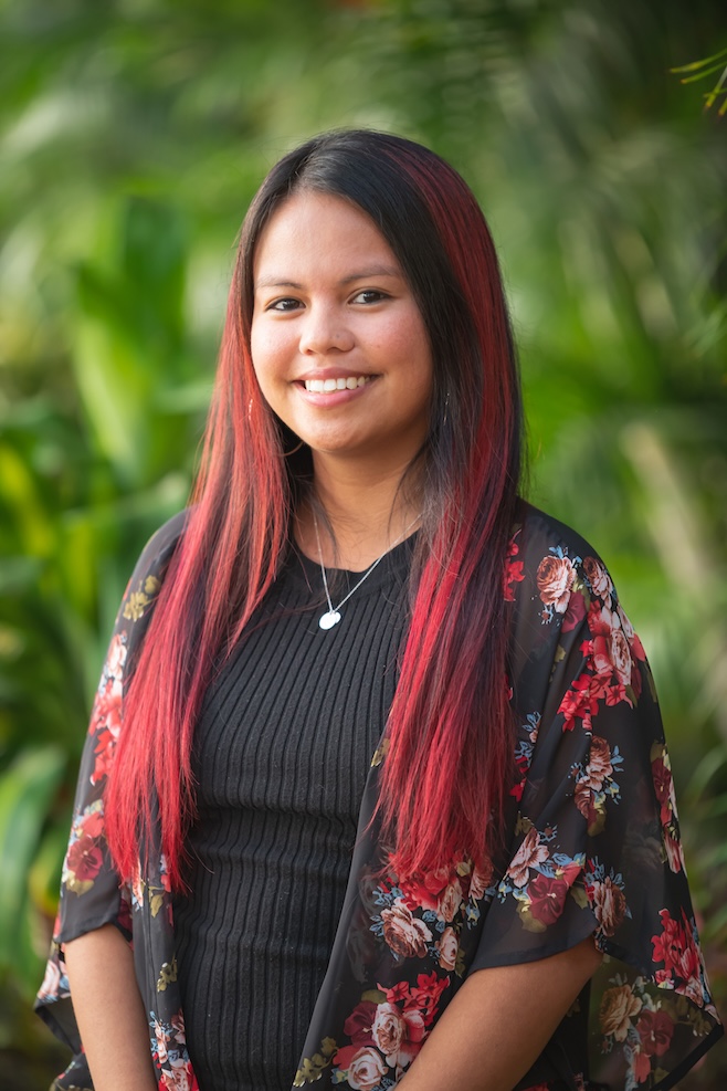 Portrait of a smiling young woman with long red-tinted hair, wearing a black ribbed top and floral kimono, outdoors with greenery behind her.
